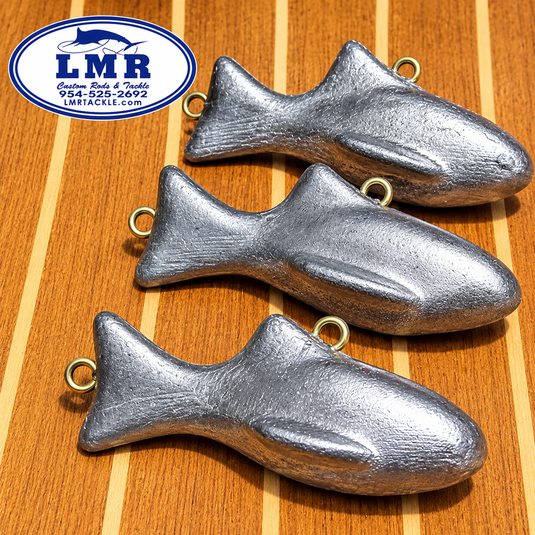 Three silver fish-shaped metal charms with gold-colored eyelets, arranged diagonally on a wooden surface. An 'LMR' brand logo is visible in the top left corner.
