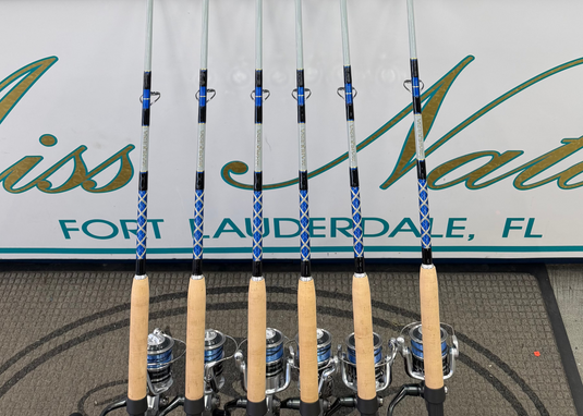 Six fishing rods with light wood handles and silver reels are lined up vertically in front of a store counter. The counter has a large sign reading 'Miss Natalie Fort Lauderdale, FL'.